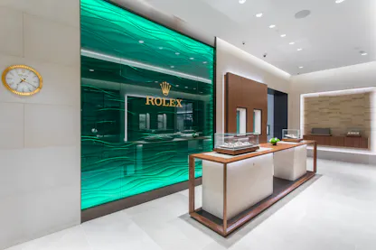 Interior shot of a Rolex store featuring a large, green, wavy glass wall with the gold Rolex logo and crown, and foreground display cases holding watches.