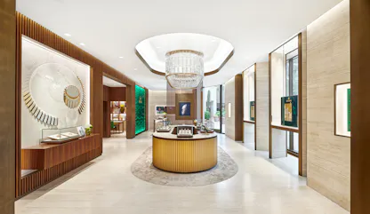 A wide shot of a modern jewelry store interior with a large, rectangular chandelier illuminating a central curved display counter. A large decorative wall feature resembling a watch mechanism is on the left, and a green backlit Rolex sign is visible in the back.
