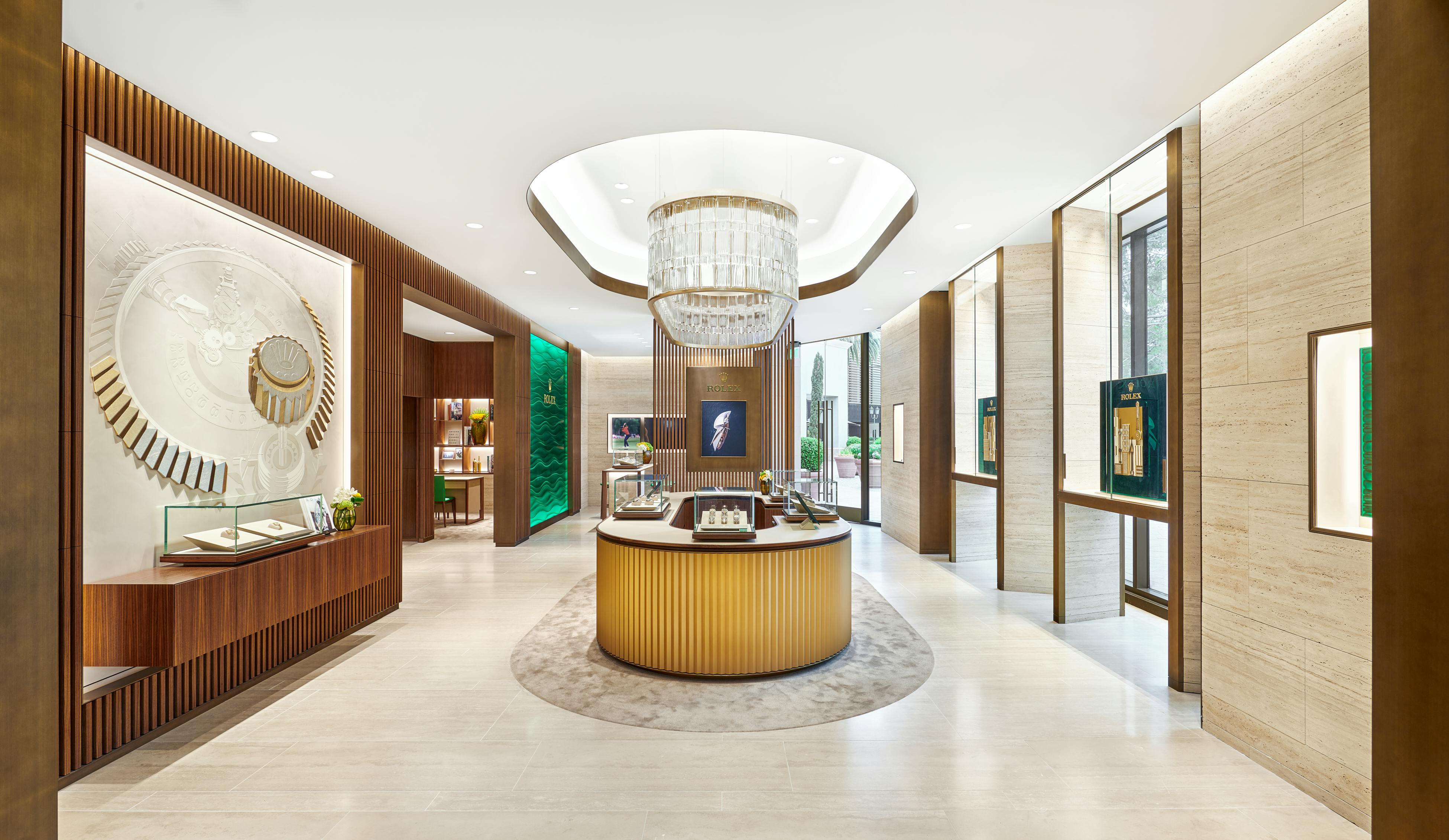 A wide shot of a modern jewelry store interior with a large, rectangular chandelier illuminating a central curved display counter. A large decorative wall feature resembling a watch mechanism is on the left, and a green backlit Rolex sign is visible in the back.