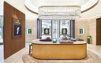 A brightly lit jewelry store interior featuring a large, rectangular chandelier over a central counter with display cases. A wooden slatted wall on the left has a Rolex display, and large windows look out onto an outdoor area.