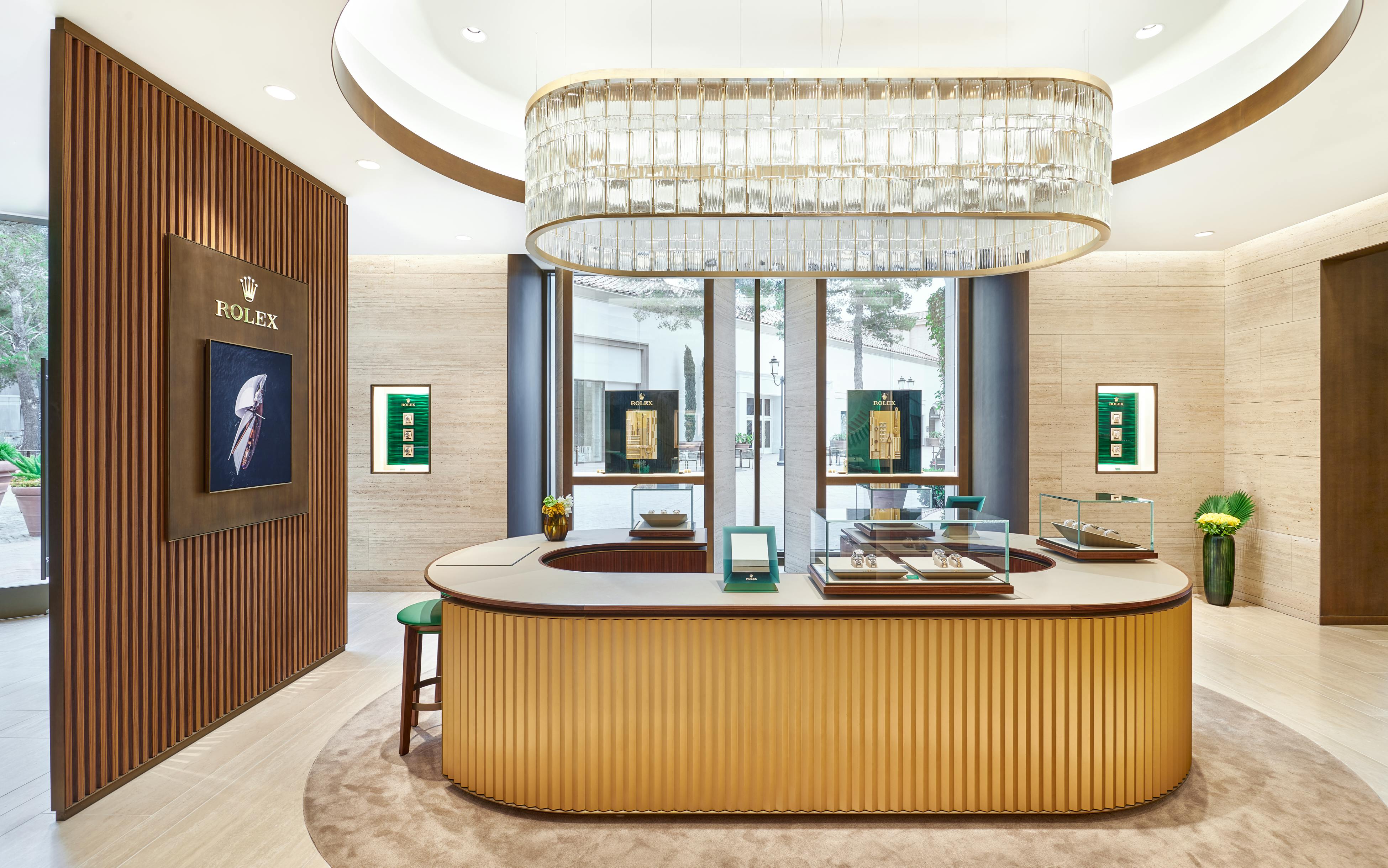 A brightly lit jewelry store interior featuring a large, rectangular chandelier over a central counter with display cases. A wooden slatted wall on the left has a Rolex display, and large windows look out onto an outdoor area.
