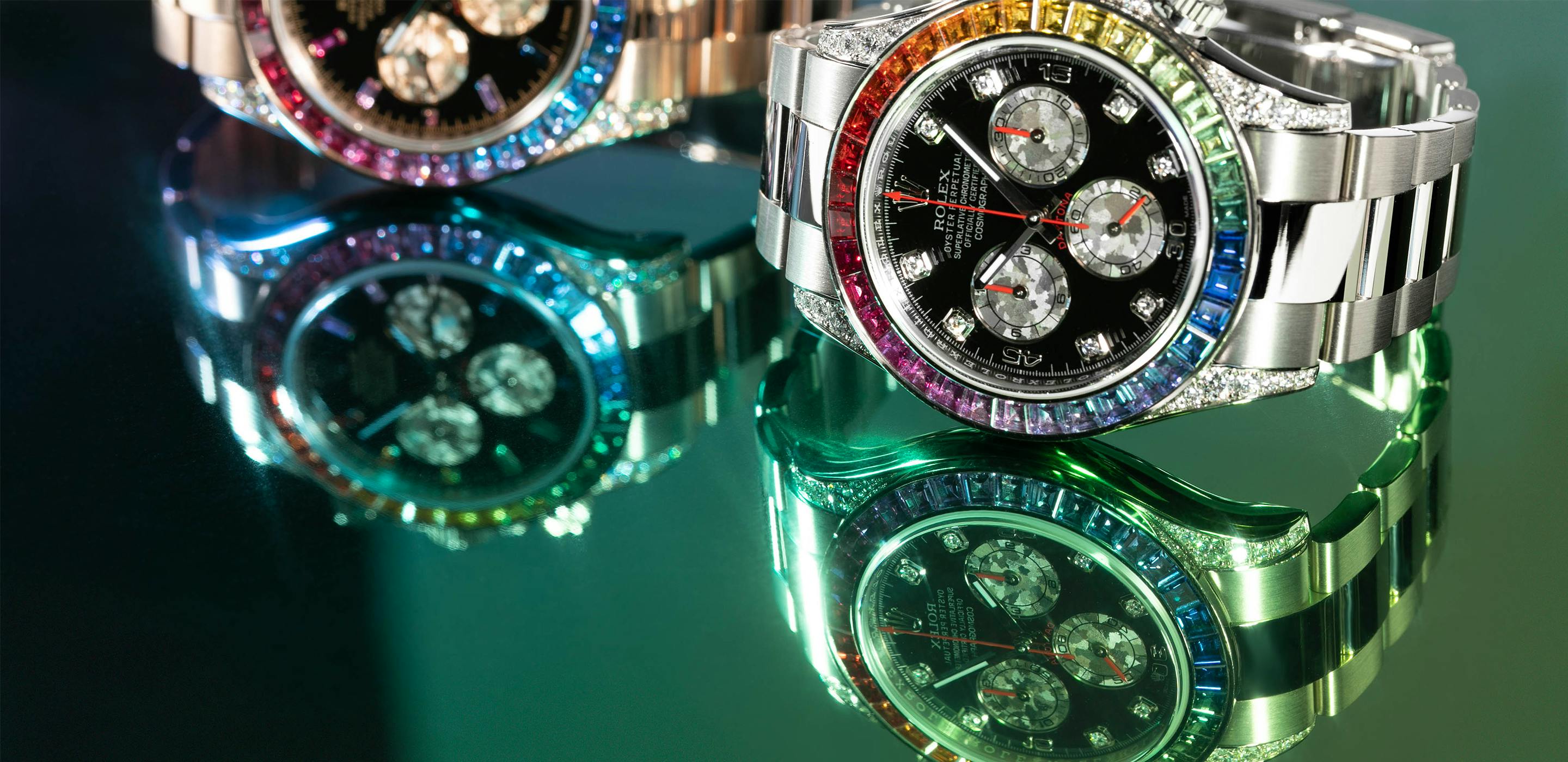 Two Rolex Rainbow Daytona watches— one in White Gold, one in Everose Gold—  sit atop a reflective green surface.