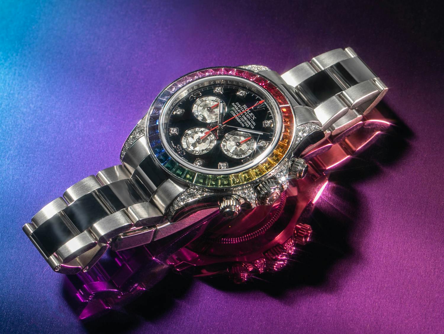 The Rolex Rainbow Daytona chronograph watch shimmers atop a blue and purple backdrop.