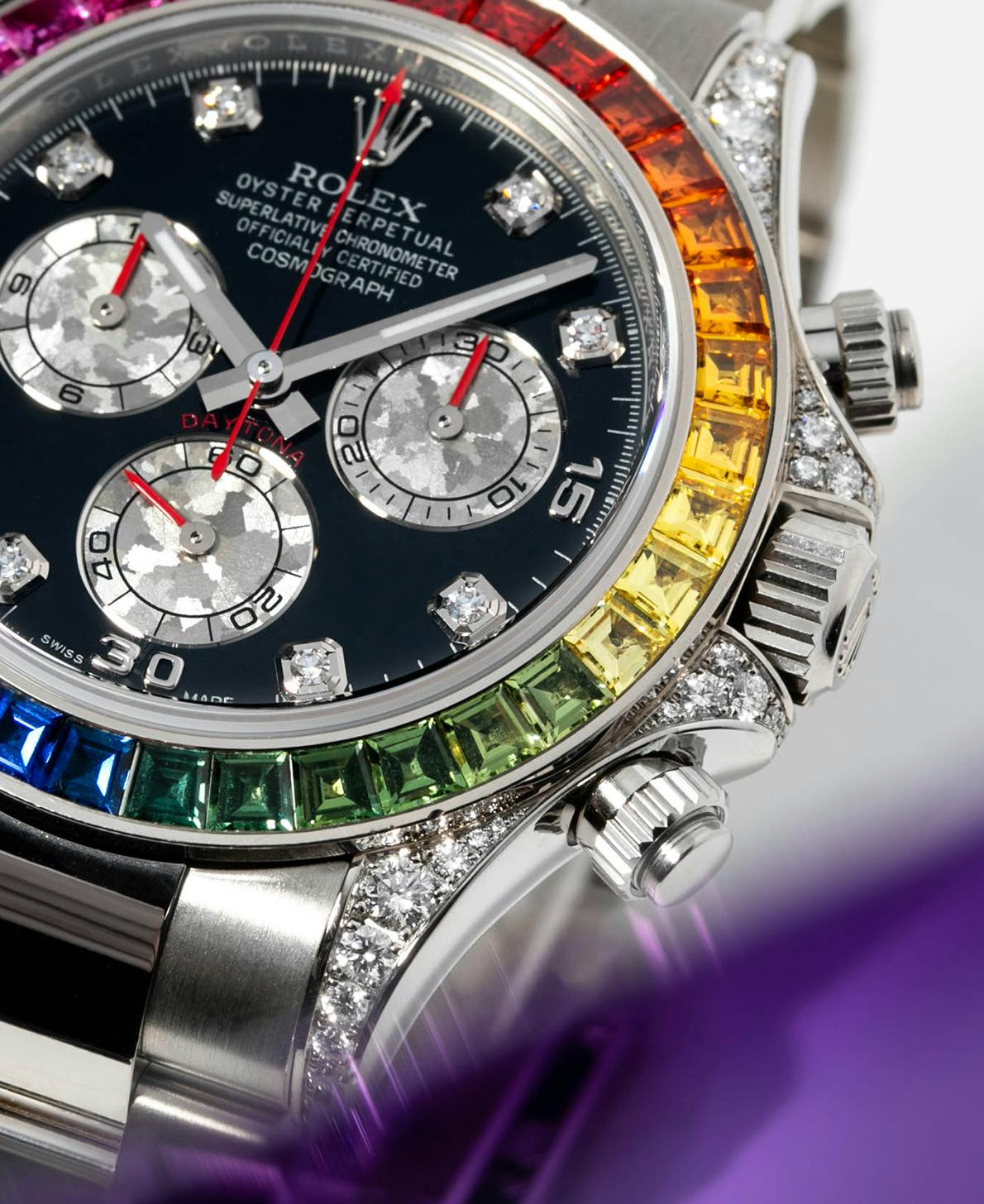 A close-up shot of the rainbow-colored gem bezel of the Rainbow Rolex Daytona watch in White Gold on a white backdrop with purple shading.